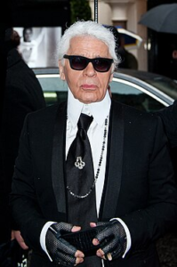 Karl Lagerfeld, who is an older man, wearing a black formal jacket, black gloves, a black tie, and black sunglasses, standing near a Fendi store.